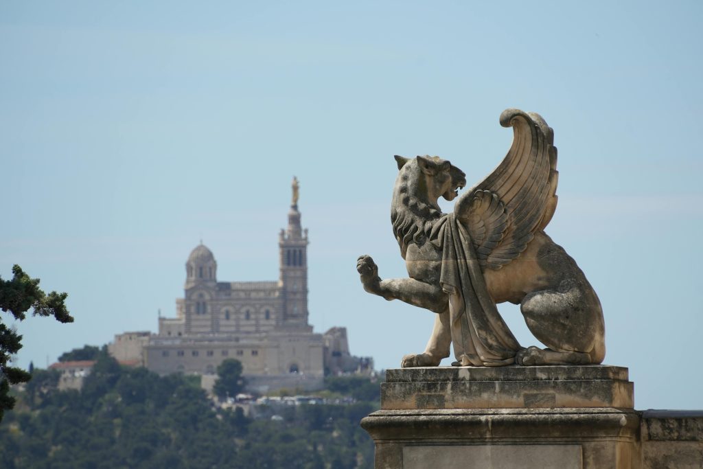 Stone sculpture with Notre-Dame de la Garde in the background, Marseille, summer sunlight.