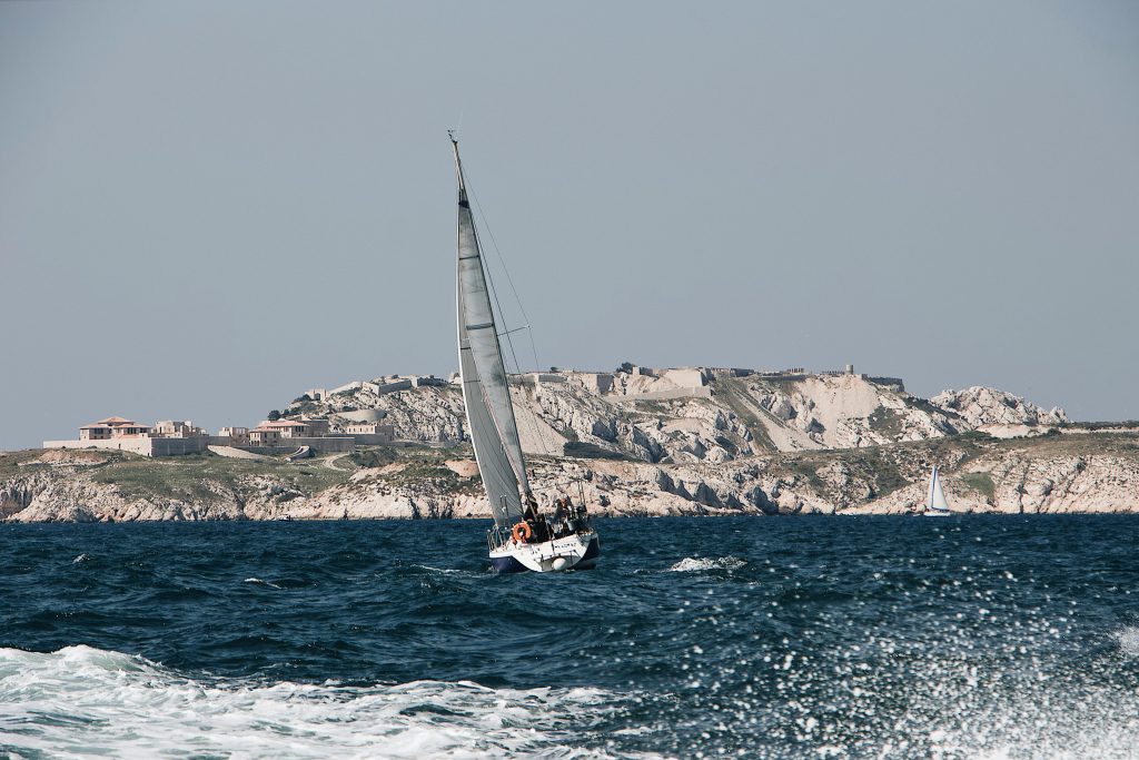 A serene scene of a sailboat navigating choppy waters near a rocky coastline with historical buildings.